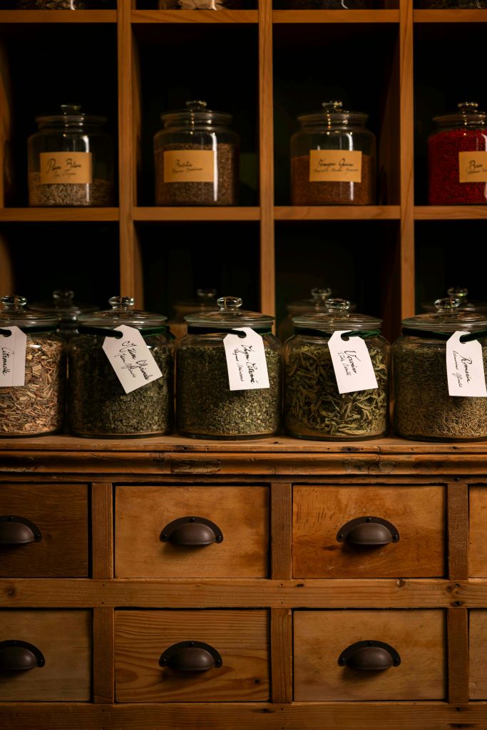 태그가 부착된 다양한 허브로 채워진 유리 항아리는 나무 선반과 조리대에 따뜻한 조명으로 배치되어 있습니다.