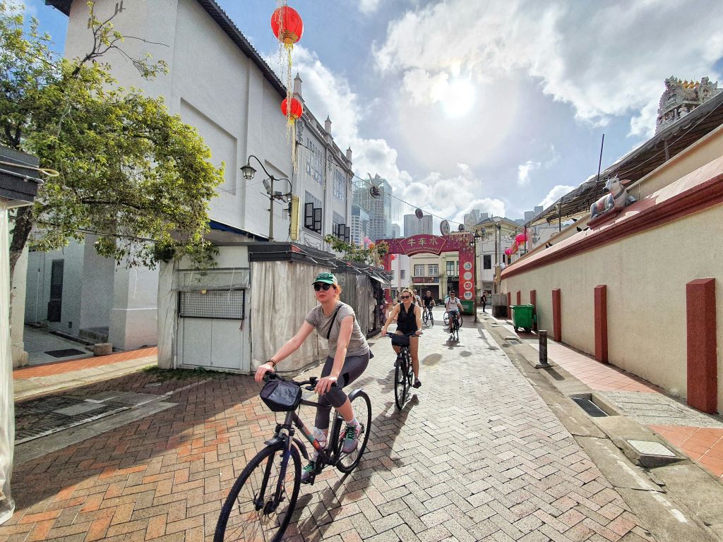 건물과 붉은 등불 장식이 늘어선 햇살 가득한 거리에서 자전거를 타는 사람들.