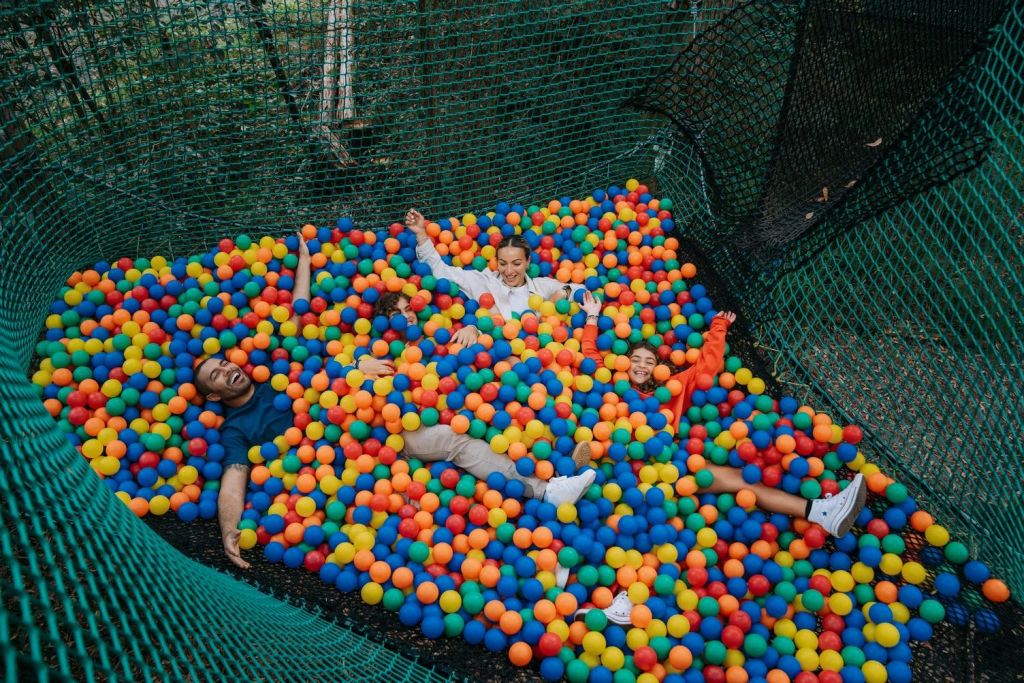 야외에서 알록달록한 플라스틱 공이 가득한 커다란 그물에서 미소 짓고 노는 사람들이 여럿 있습니다.