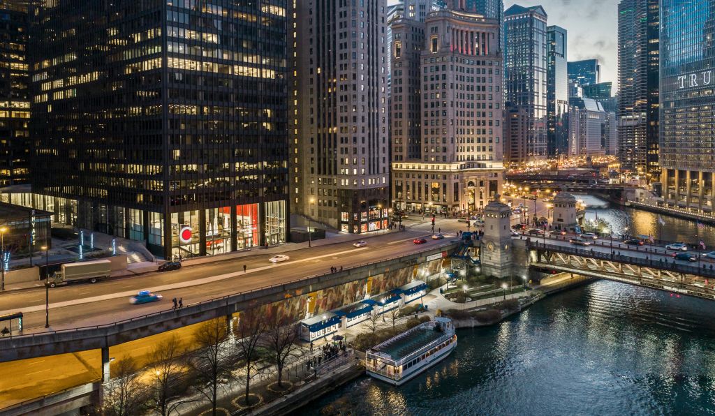 높은 건물이 있는 도시 풍경, 자동차가 있는 번화한 도로, 보트가 있는 바닷가, 강둑을 따라 걷는 보행자.