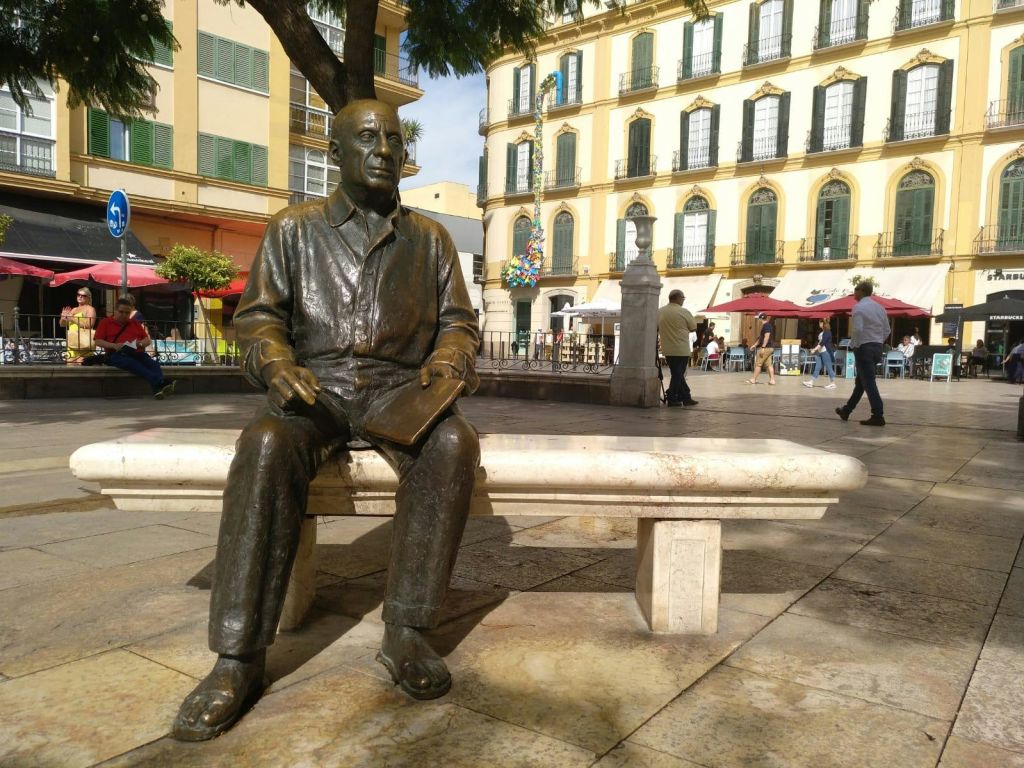 건물, 사람, 나무가 배경으로 펼쳐진 번화한 광장의 벤치에 앉아 있는 한 남자의 청동 동상.