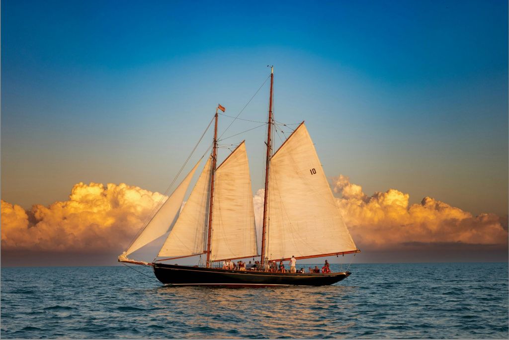 황금빛 구름과 푸른 하늘을 배경으로 잔잔한 물 위에 두 척의 커다란 돛이 달린 요트.