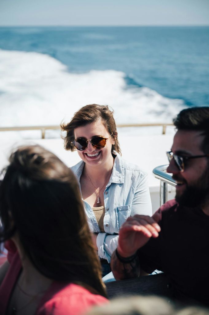 세 사람이 보트 타기를 즐기고 있으며, 두 사람은 미소를 지으며 선글라스를 끼고 있습니다. 배경에는 바다가 보입니다.