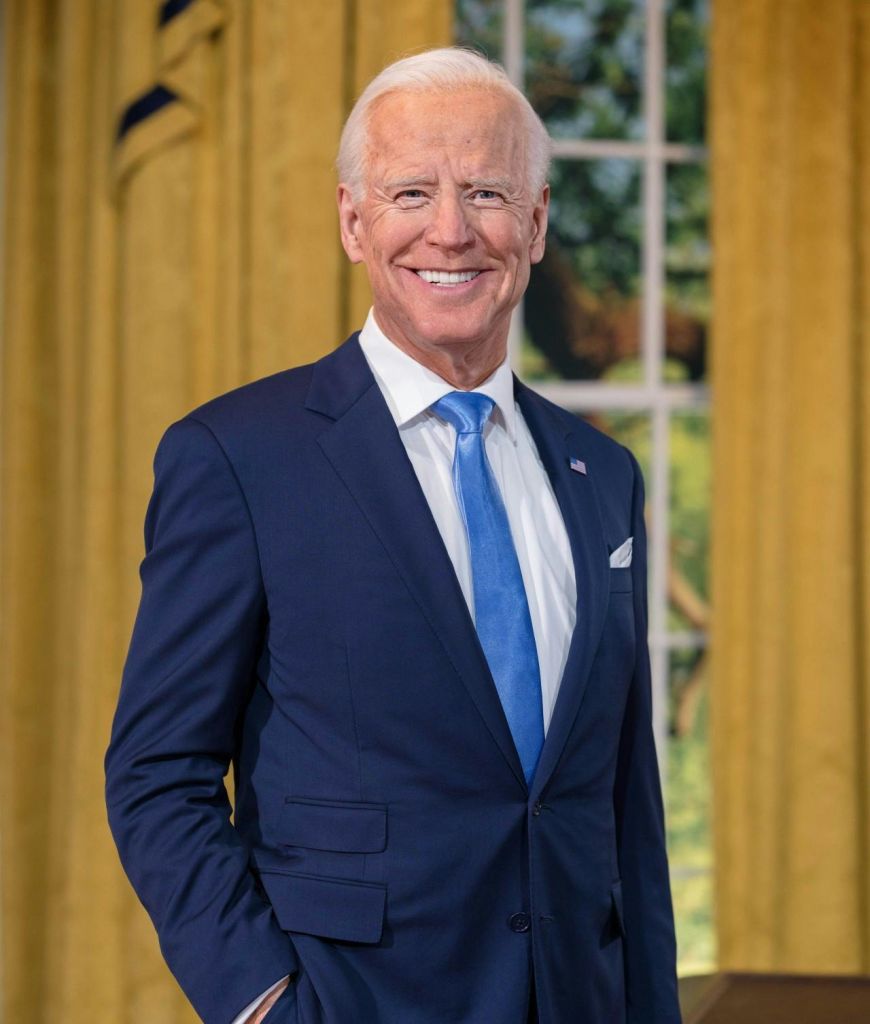 파란색 정장을 입고 파란색 넥타이를 매고 노란색 커튼과 외부의 녹지가 보이는 창문 앞에 서서 웃고 있는 남성.