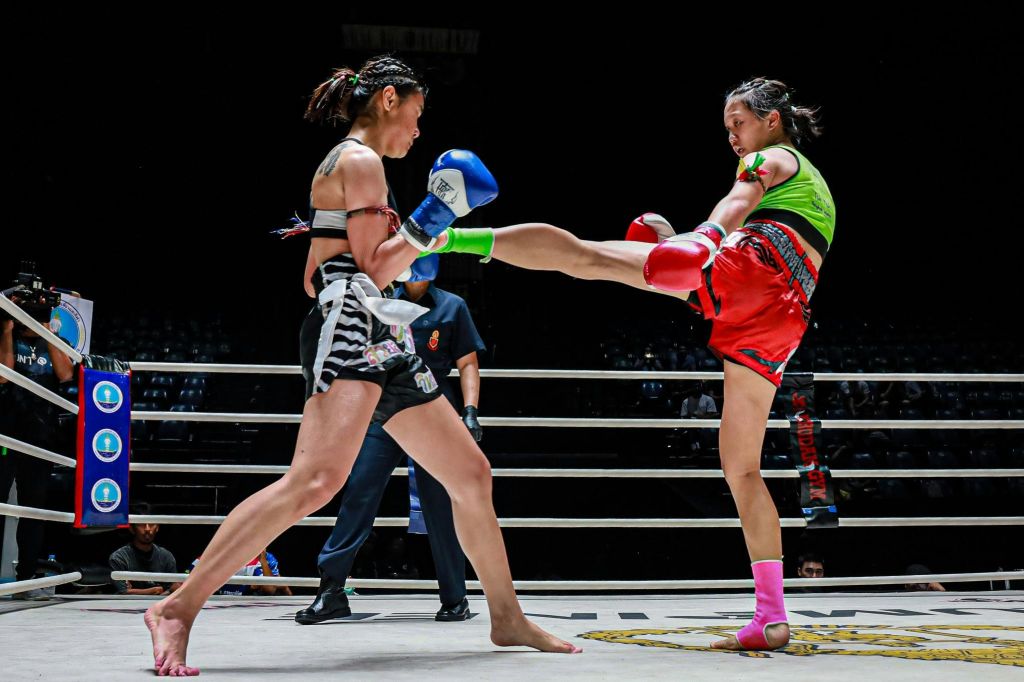 한 반지에 두 명의 무에타이 여성 선수가 있고, 한 명은 하이킥을 내고 있습니다. 둘 다 장갑과 운동 장비를 착용합니다. 백그라운드에서 심판.