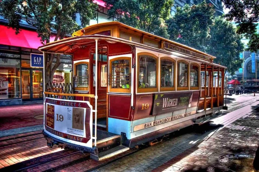 나무와 건물 옆에 19라는 숫자가 있는 빨간색과 노란색의 샌프란시스코 케이블카로, 뮤지컬 '위키드' 를 홍보합니다.