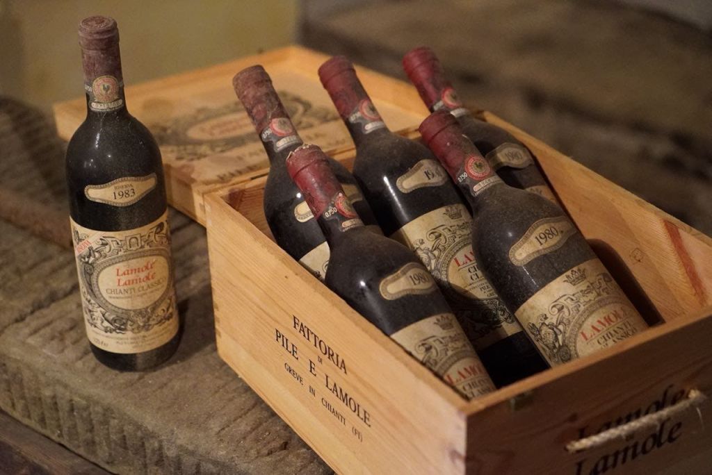 "Fattoria Pile e Lamole" 이라고 적힌 나무 상자에 여섯 개의 먼지투성이 와인병이 놓여 있고, 그 옆에 한 개의 서 있는 병이 있습니다.