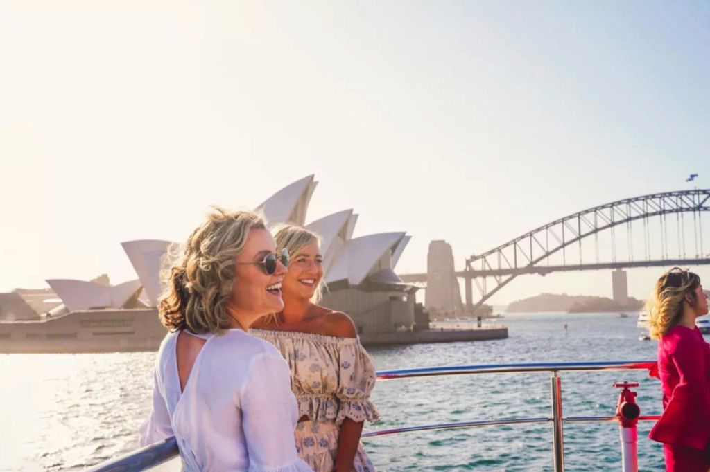 화창한 날 시드니 오페라하우스와 하버 브리지를 배경으로 한 보트에서 웃고 있는 두 사람.