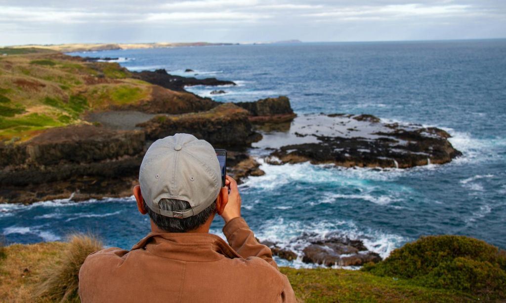바위가 많은 해안선과 바다 전망을 배경으로 회색 모자를 쓴 사람이 사진을 찍고 있습니다.