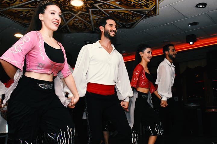 화려한 천장 조명 아래에서 전통 무용복을 입은 네 명이 손을 잡고 미소를 짓고 있습니다.