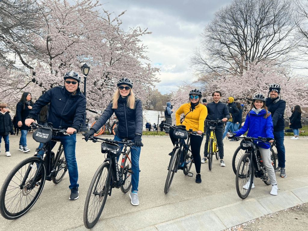자전거를 탄 세 사람이 헬멧을 쓰고 벚꽃이 만발한 길에서 포즈를 취하고 있습니다. 다른 사람들은 백그라운드에서 걷습니다.