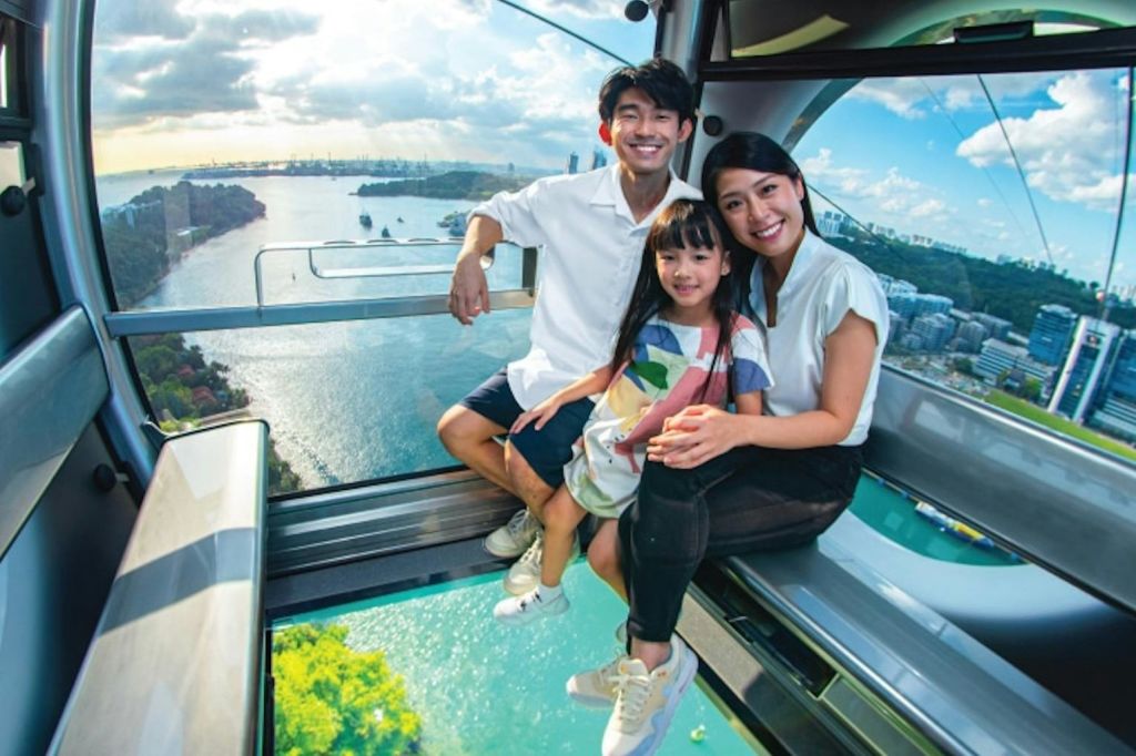 유리 케이블카 안에는 물, 보트, 도시 건물이 한눈에 내려다보이는 아름다운 풍경을 배경으로 웃고 있는 세 가족이 앉아 있습니다.