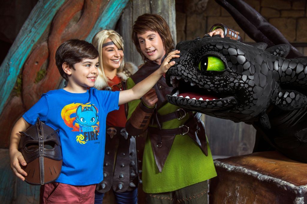 파란색 셔츠를 입은 한 소년이 중세 의상을 입은 두 명의 성인과 함께 드래곤 헤드 소품을 만지며 모두 웃고 있습니다.