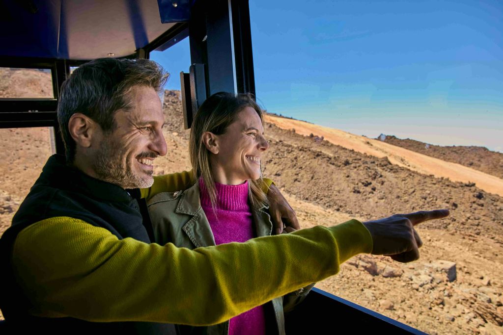두 사람이 미소를 지으며 차량 창밖을 내다보고 있으며, 바깥에는 건조하고 언덕이 많은 풍경이 보입니다.