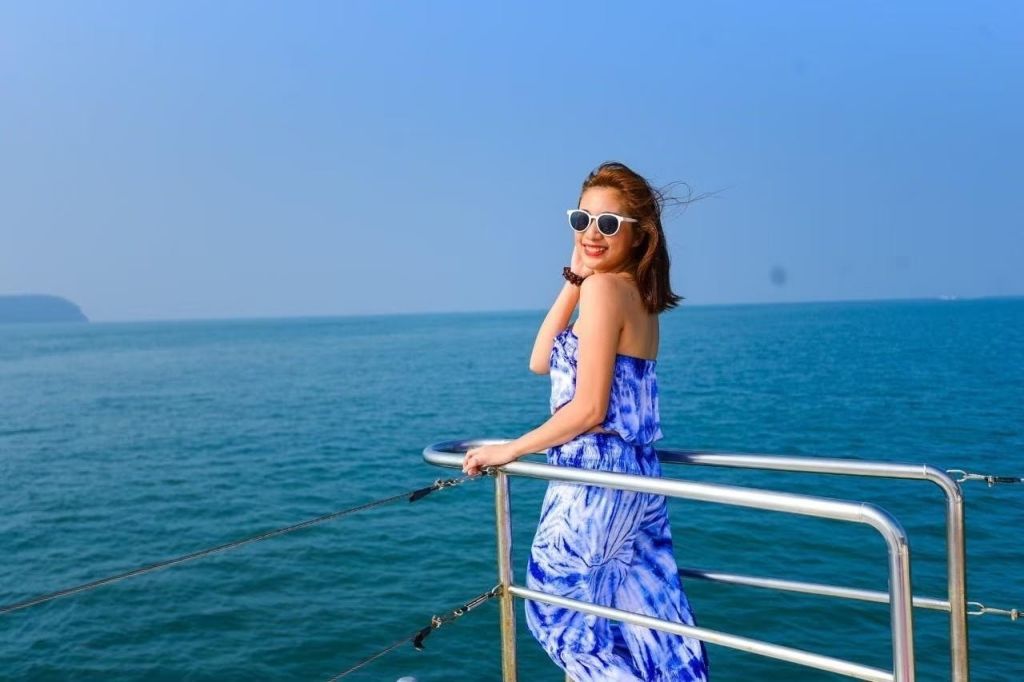 파란색 드레스와 흰색 선글라스를 쓴 여성이 바다와 맑고 푸른 하늘을 배경으로 보트 난간에서 미소를 짓고 있습니다.