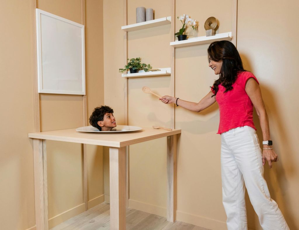 한 소년의 머리가 테이블의 구멍을 뚫고 나타나고, 한 여성이 나무 숟가락을 들고 놀리듯 근처에 서 있습니다.