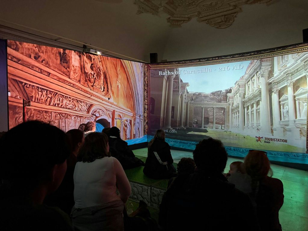 한 무리의 사람들이 방에 앉아 역사 건축물을 화면에 투영하는 모습을 지켜보고 있습니다. 디스플레이에 "Baths of Caracalla - 216 AD" 라는 텍스트가 표시됩니다.