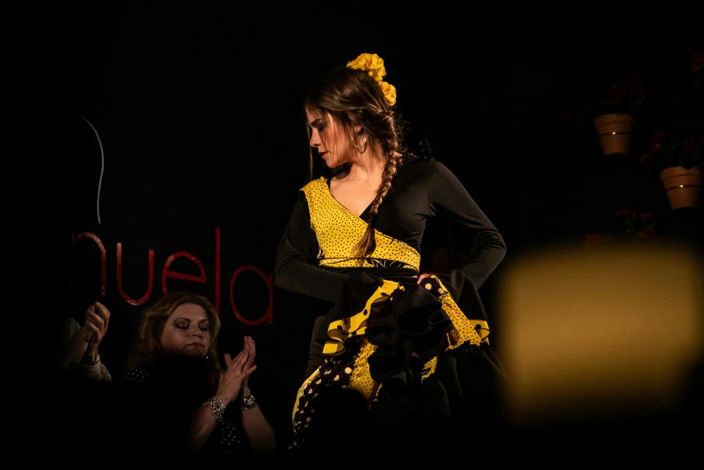 노란색과 검은색 드레스를 입은 공연자가 무대에 서고, 다른 사람이 배경에서 희미한 조명 아래 박수를 치고 있습니다.