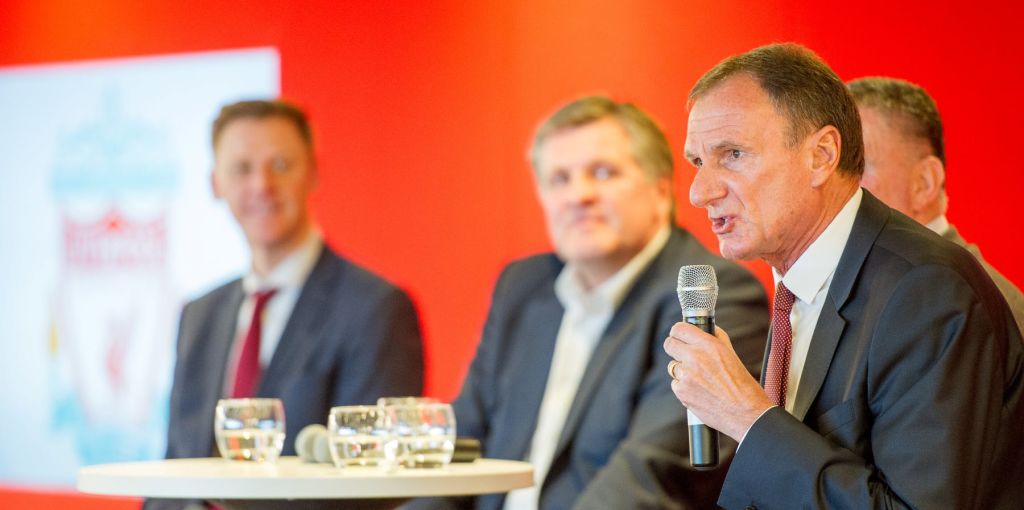 양복을 입은 남성이 다른 남성이 등장하는 흐릿한 배경과 빨간색 벽이 있는 마이크를 향해 말을 걸고 있습니다.