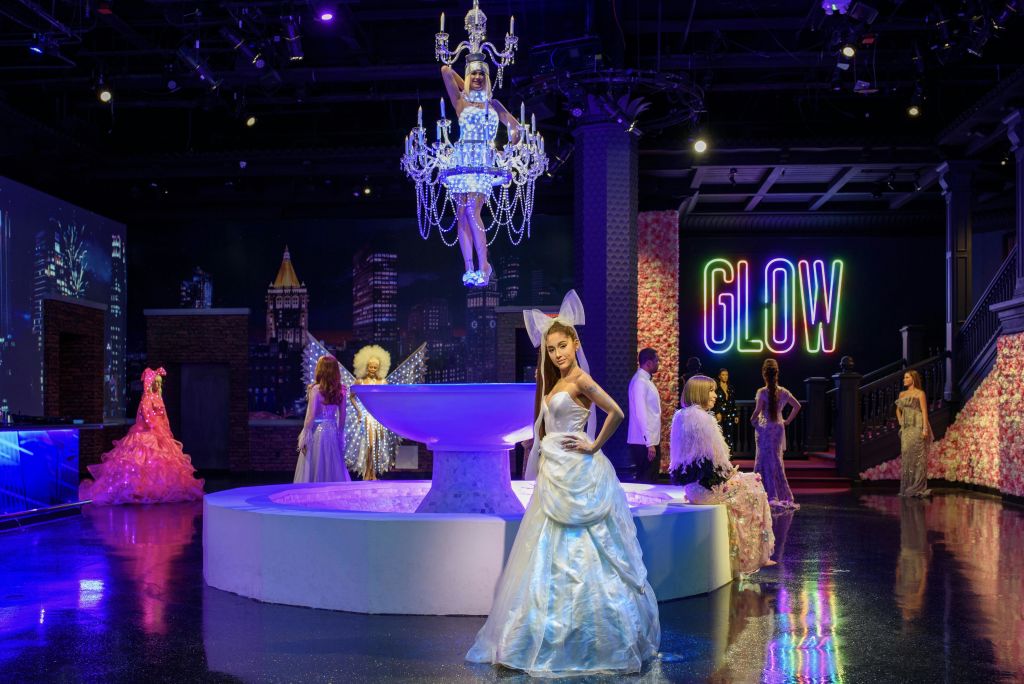 밝은 파란색 가운을 입은 왁스 피규어가 분수대 근처에 서 있으며, 네온 "야광" 표지판 아래 샹들리에에 매달린 다른 피규어가 있습니다.