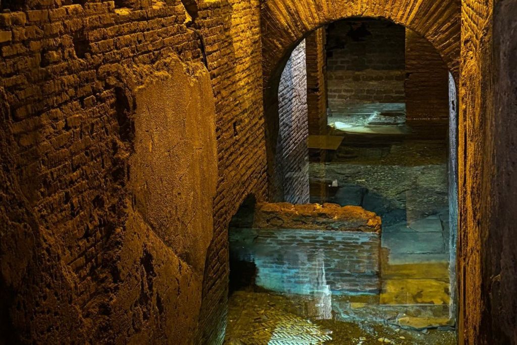 벽돌 벽과 아치, 바닥에 반사되는 수면이 있는 조명이 희미한 지하 복도입니다.