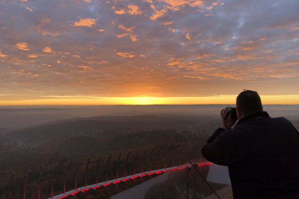 한 사람이 광활한 풍경이 내려다보이는 경치 좋은 석양에 난간과 빛나는 붉은 빛을 배경으로 쌍안경을 들여다보고 있습니다.