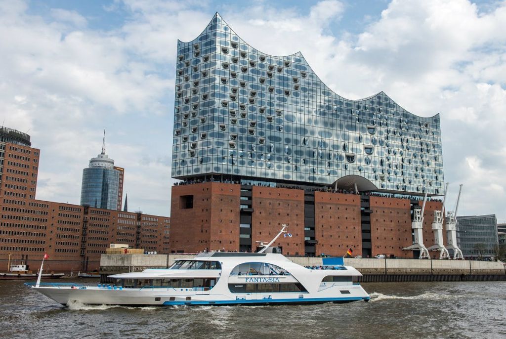 일부 흐린 하늘 아래 벽돌 받침이 있는 커다란 파도 모양의 유리 건물인 엘프필하모니 함부르크 (Elbphilharmonie Hamburg) 를 지나 현대적인 보트 항해.