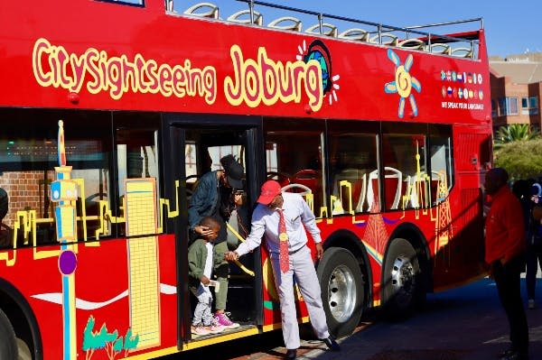 빨간색 관광 버스에는 "Joburg" 라는 라벨이 붙어 있으며, 버스에서 내리는 어린이와 성인을 돕는 가이드가 있습니다.