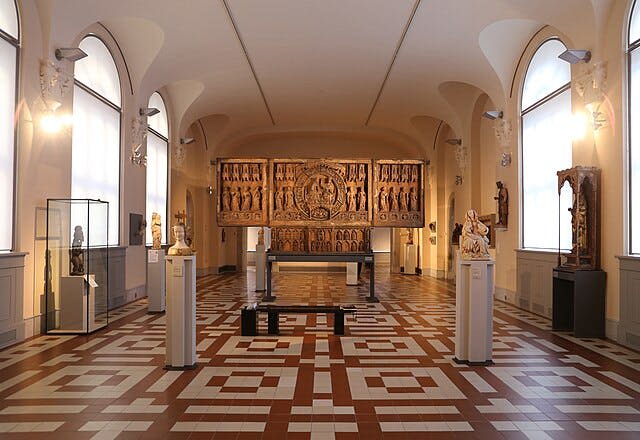 받침대에 동상이 있는 넓은 박물관 갤러리, 뒷벽에 정교한 목조 예술품, 기하학적 타일 바닥이 있습니다.