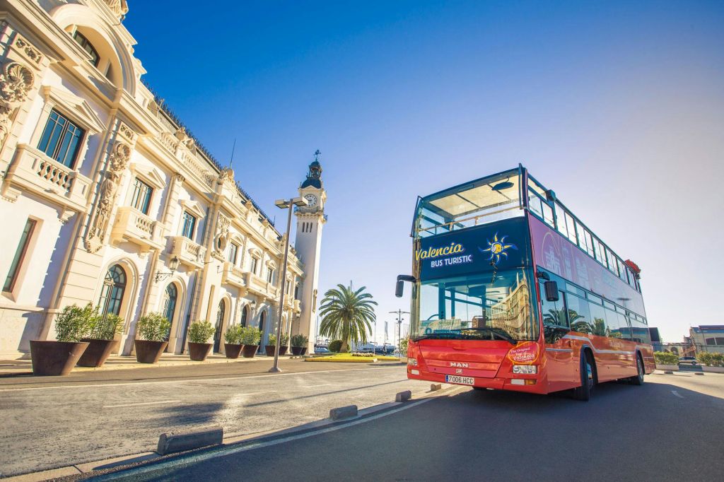 "Valencia Bus Turistic" 이라는 이름의 빨간색 이층 투어 버스가 화창한 날 유서 깊은 흰색 건물과 야자수 옆에 주차되어 있습니다.