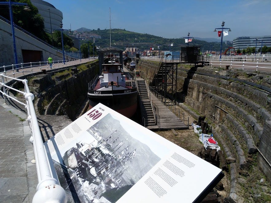 맑은 하늘 아래 건물과 언덕을 배경으로 두 척의 보트가 있는 마른 부두가 안내판에 내려다보입니다.