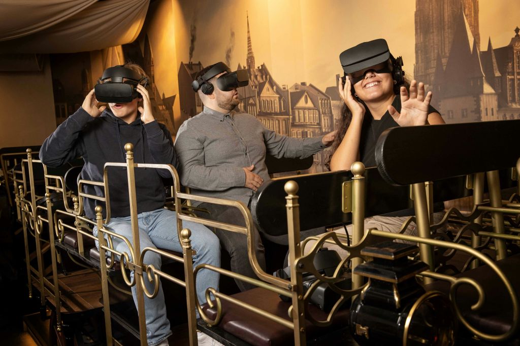 VR 헤드셋을 착용한 두 사람이 화려한 레일과 함께 차량에 앉아 가상 환경과 상호 작용하는 것처럼 웃으며 손을 내밀고 있습니다.