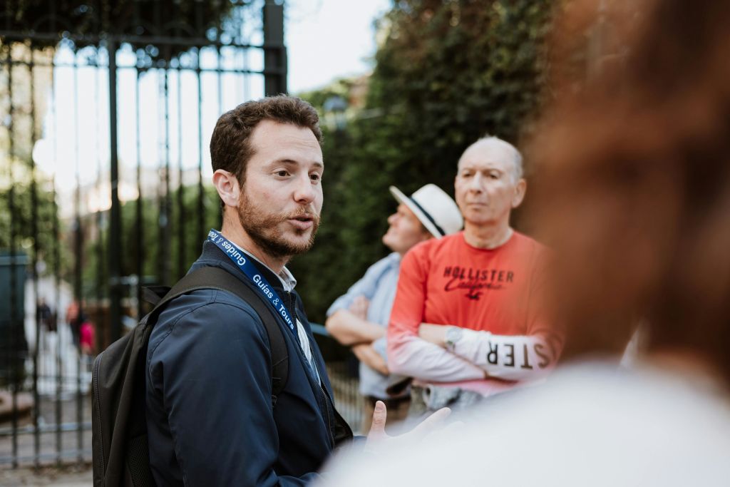 투어 가이드가 야외에서 여러 사람들과 이야기를 나눕니다. 빨간 셔츠를 입은 한 명과 모자를 쓴 다른 한 명은 배경에서 주의 깊게 경청합니다.