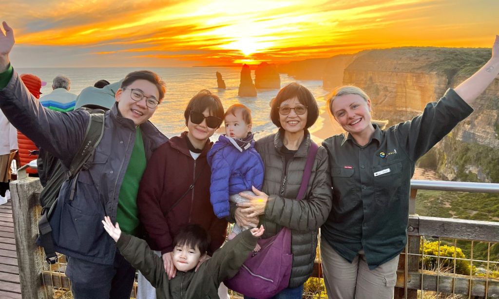 바위 지층과 바다를 배경으로 석양을 바라보며 미소를 짓는 5인조 그룹. 앞에 있는 어린이가 팔을 뻗습니다.