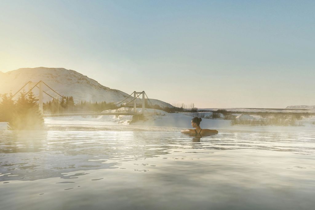 한 사람이 눈 덮인 풍경, 다리, 산을 배경으로 맑은 하늘 아래 펼쳐진 찜질방 야외 수영장에서 휴식을 취하고 있습니다.