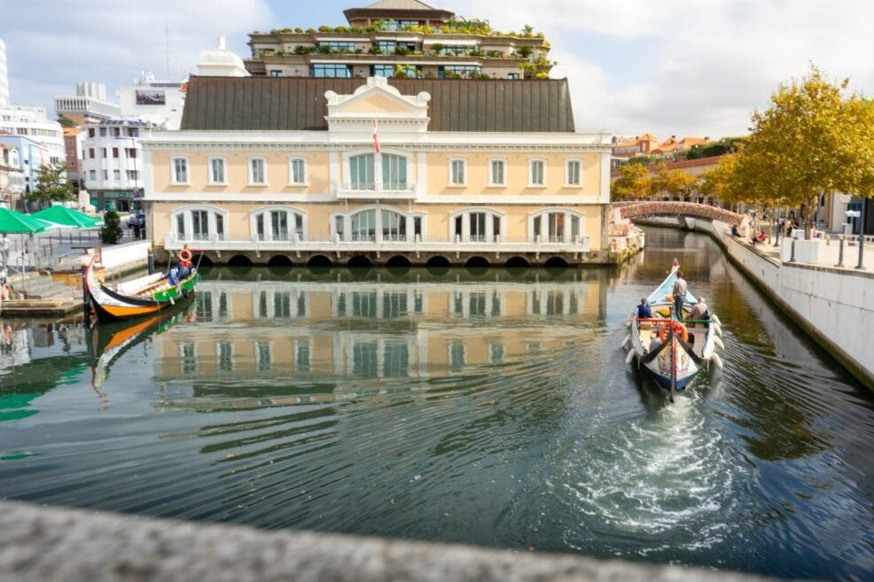아치형 창문이 있는 화려한 베이지색 건물 앞에서 두 척의 보트가 고요한 운하를 따라 이동하고 있습니다. 나무와 다른 건물들이 배경으로 보입니다.