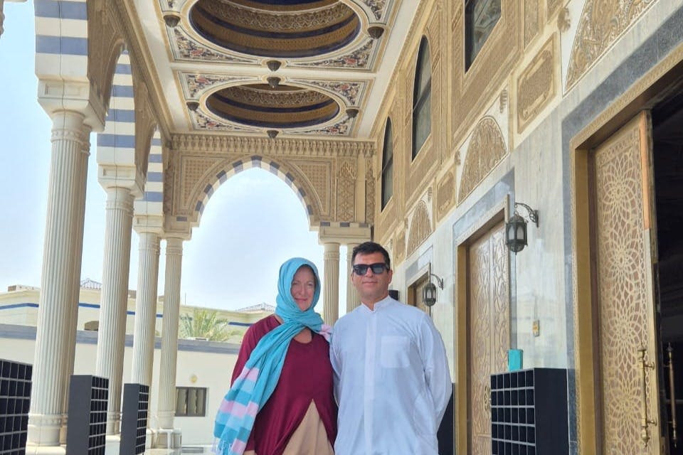 정교한 디자인과 기둥이 돋보이는 화려한 아치형 통로 아래 두 사람이 나란히 서 있습니다. 두 사람 모두 전통적인 복장을 하고 있습니다.