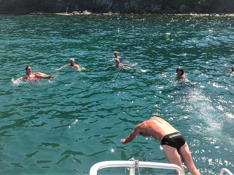 바위가 많은 해안가 근처의 맑은 물속에서 수영하는 사람이 몇 명 있으며, 한 명이 전경의 보트에서 뛰어듭니다.