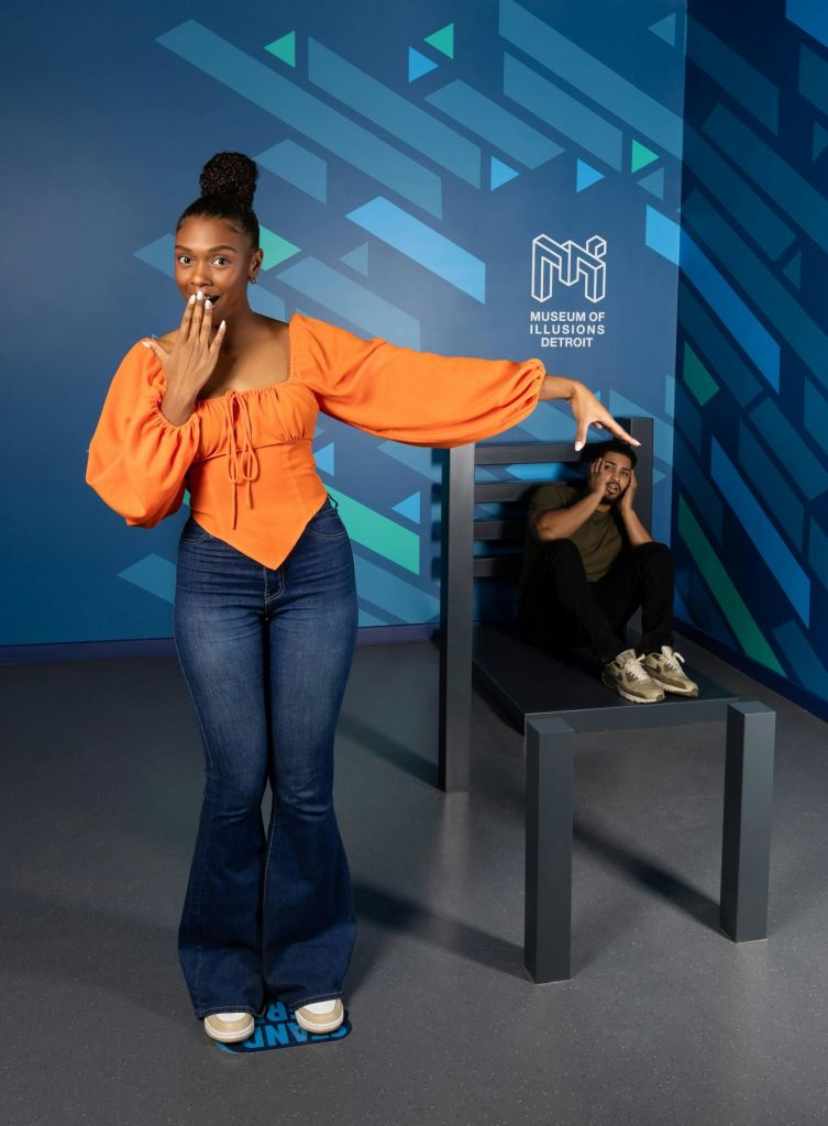 주황색 상의와 청바지를 입은 여성이 디트로이트 환상 박물관의 의자에 앉아 있는 남성을 향해 장난스럽게 제스처를 취하고 있습니다.