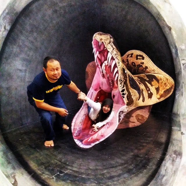 한 남성이 커다랗고 입이 열린 뱀 조각상 안에서 장난스럽게 포즈를 취하는 여성을 돕고 있습니다.