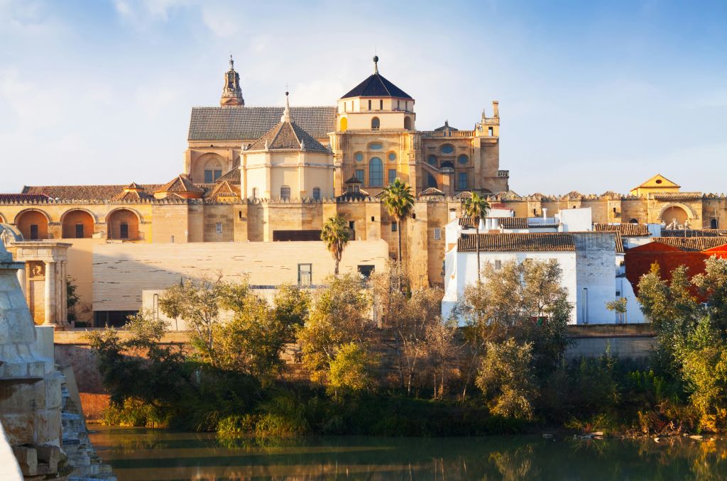 맑고 푸른 하늘 아래 나무와 강이 줄지어 있는 돔형 건물과 첨탑 모양의 지붕이 있는 역사적인 건물입니다.