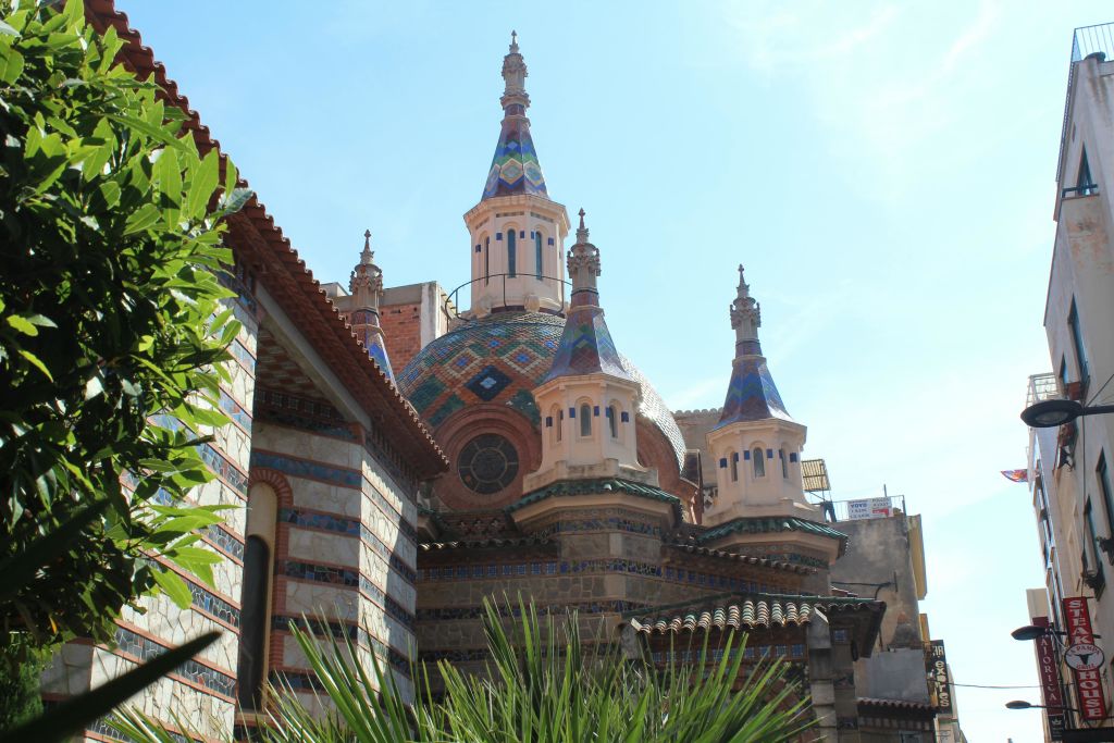 형형색색의 타일 돔과 화려한 첨탑이 있는 건물로, 맑은 하늘 아래 식물과 부분적으로 보이는 건물로 둘러싸여 있습니다.