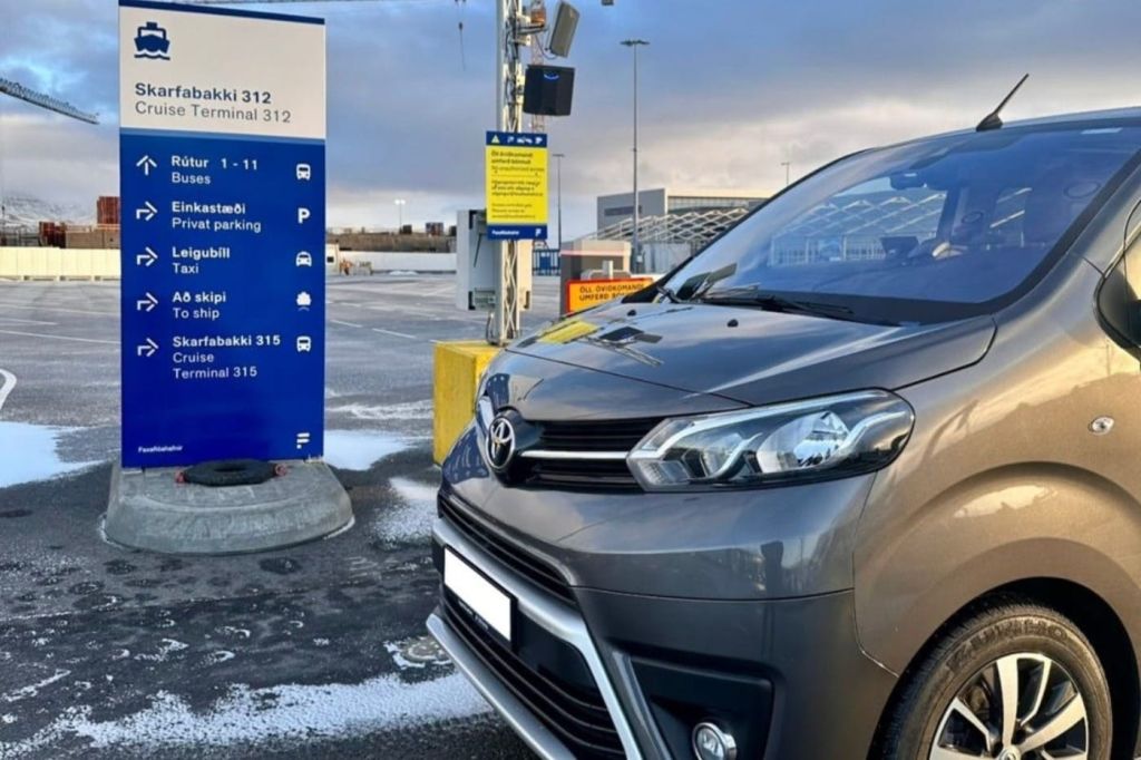 파란색과 노란색 표지판 근처에 주차장과 터미널 방향을 알리는 회색 밴이 주차되어 있습니다. 배경에 흐린 하늘.