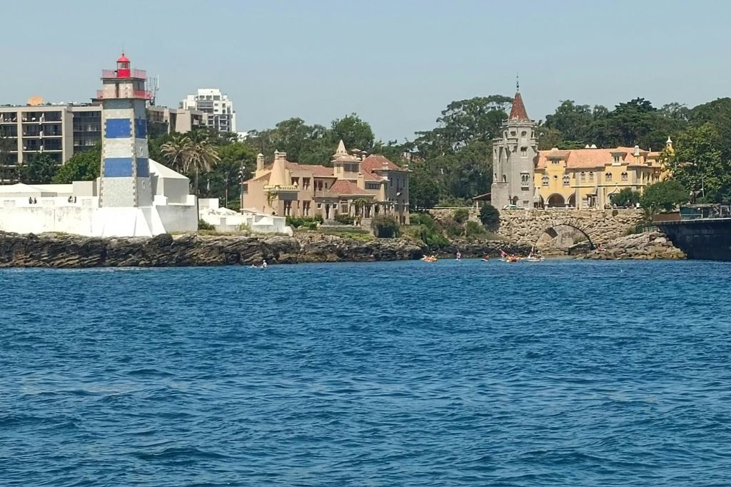 등대, 역사적인 건물, 바위가 많은 해안선이 있는 해안 풍경. 몇몇 사람들이 활기찬 푸른 바다에서 카약을 타고 있습니다.