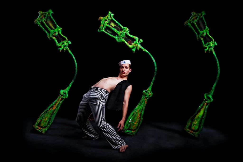 줄무늬 바지와 모자, 조끼를 입은 사람이 극적으로 몸을 뒤로 기울이고 있습니다. 세 개의 추상적인 녹색 조각이 배경에 있습니다.