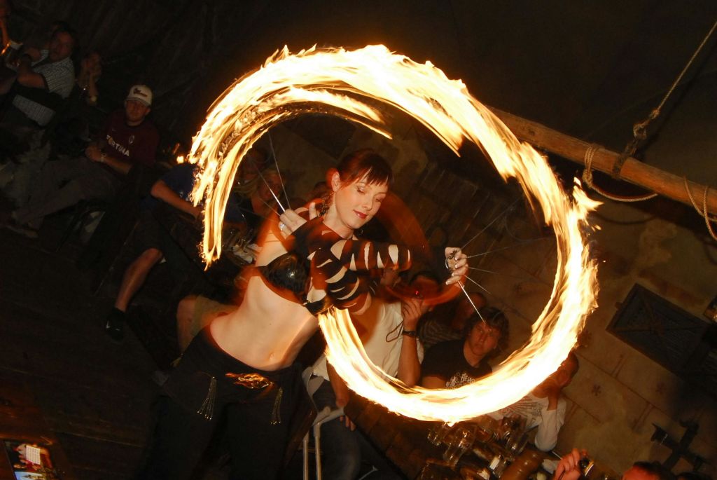 조명이 어두운 방에서 커다란 불꽃 후프와 함께 불춤을 추는 여성, 구경꾼들이 배경을 지켜보는 모습.