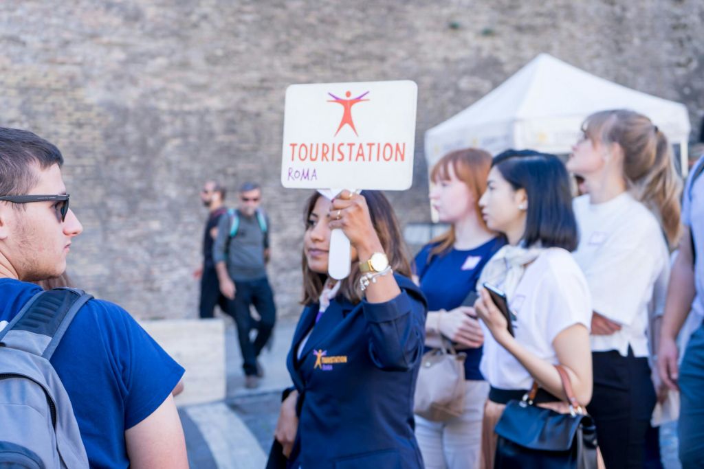 네이비 블레이저를 입은 투어 가이드가 "Touristation Roma" 표지판을 들고 있어 세심한 관광객들을 역사적인 배경으로 안내합니다.