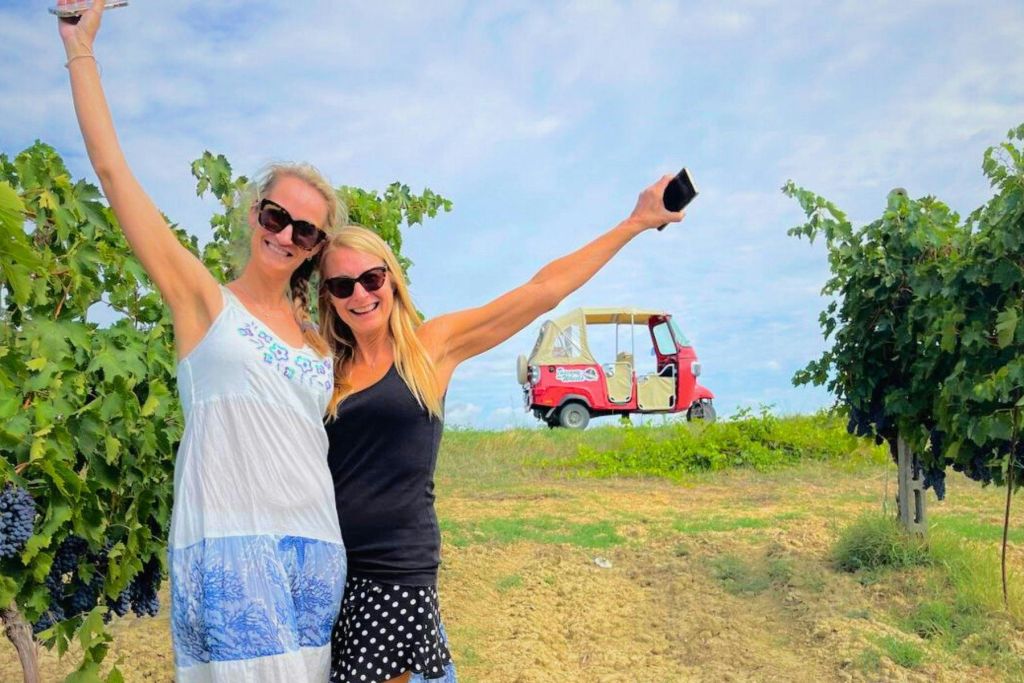 포도밭에서 미소를 짓고 포즈를 취하고 있는 두 명의 여성이 포도나무를 배경으로 작은 빨간색 차량을 배경으로 포즈를 취하고 있습니다.