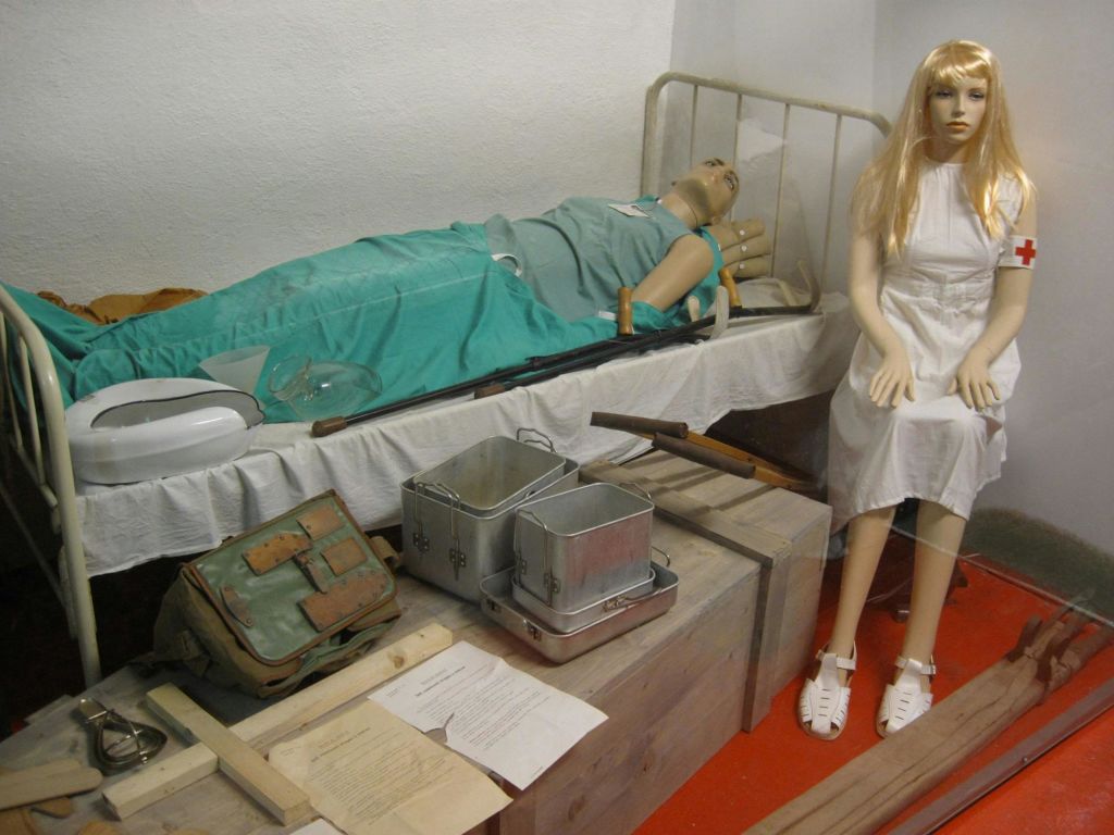 빈티지 의료 마네킹을 묘사하는 장면: 한 명은 상자에 앉아있는 간호사로 옷을 입고 다른 한 명은 병원 침대에 앉아 있습니다. 근처의 의료 용품.