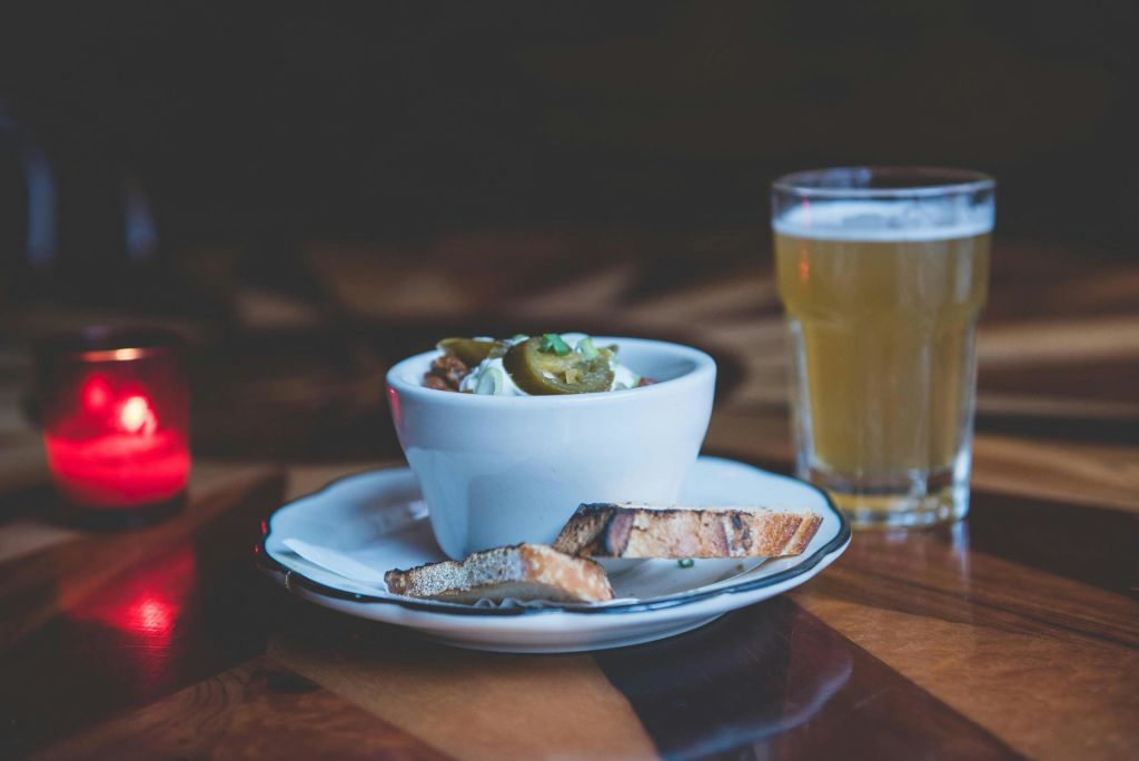 나무 테이블 위에 놓인 맥주 한 잔 옆에는 할라피뇨를 얹은 음식이 담긴 빵 조각이 담긴 하얀 그릇이 접시에 담겨 있습니다.